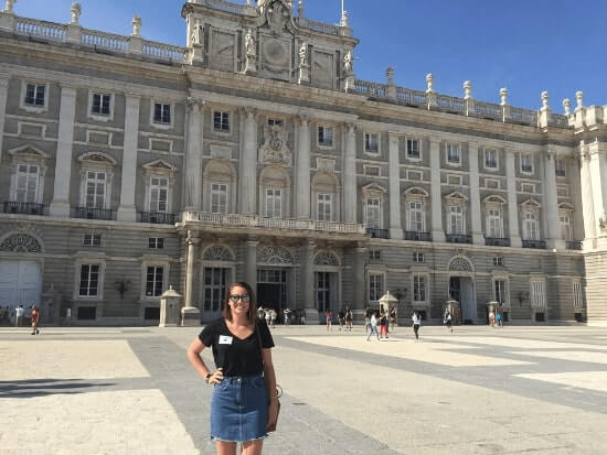 A woman stands smiling in front of a large historic palace building on a sunny day