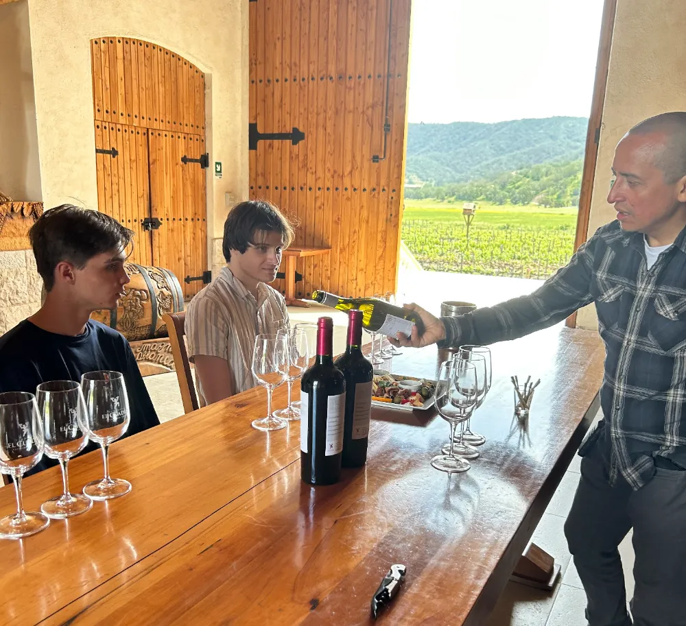 Man pouring wine for two people at a tasting table inside a vineyard building