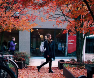 Student walking past a building along a path lined with orange autumn leaves.