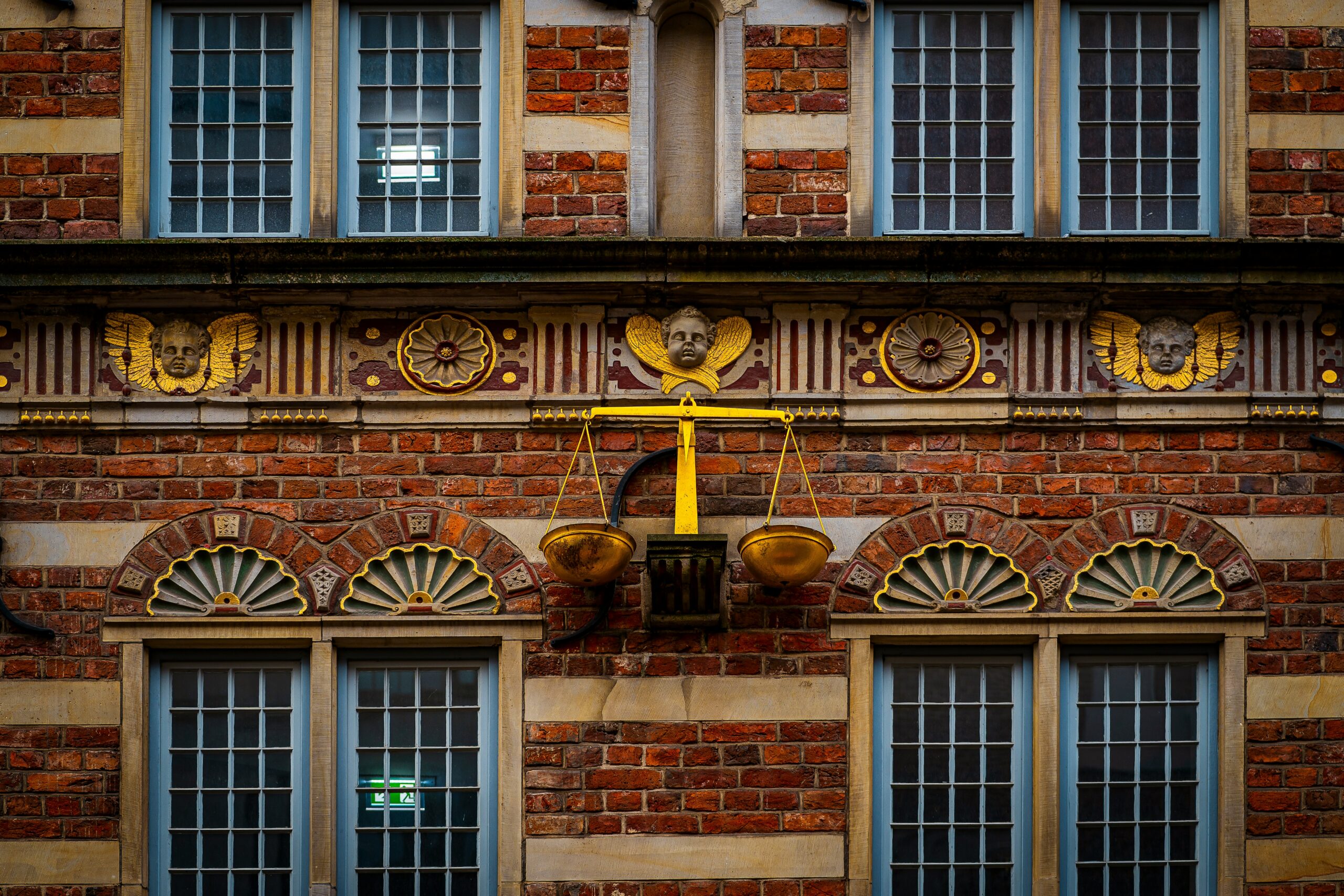 An ornate red brick building facade featuring golden scales of justice mounted between grid windows and carved cherub details.