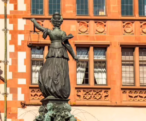 Rear view of a bronze Lady Justice statue holding scales aloft, set against a red sandstone historic building.