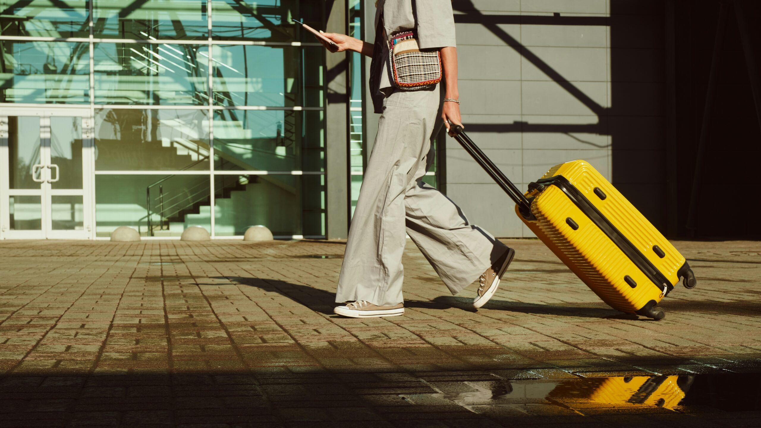 Traveler walking outside a modern airport terminal holding a phone and pulling a suitcase